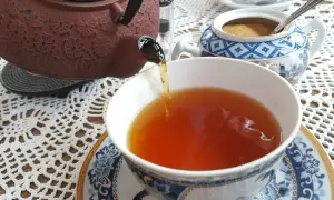 Ciemnobrązowy kolor herbaty w filiżance, nalewanej z ozdobnego czajniczka. Obok cukiernica z brązowym cukrem.
